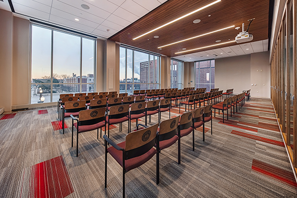 The room features rows of brown, cushioned chairs with wooden backs. The ceiling has built-in lighting, and a projector is installed. Large windows on the left side allow natural light, presenting a view of buildings outside. The right side has glass-paneled walls. The carpet is primarily gray with red accents.