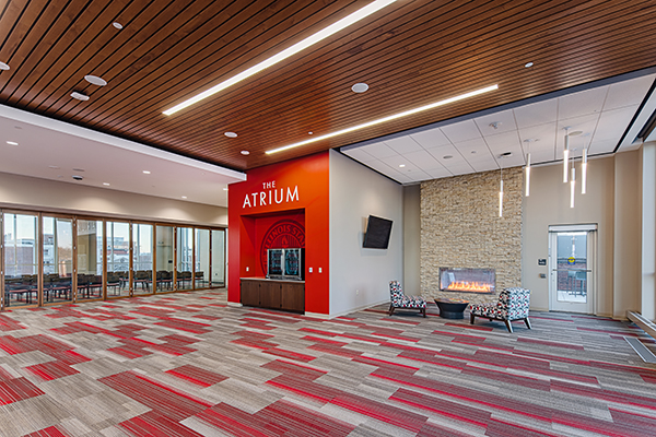 The floor is covered with a red and gray patterned carpet, creating a dynamic visual effect. The ceiling is wood-paneled with recessed lighting strips. A prominent red wall on the left side displays the text THE ATRIUM in white. Adjacent to this wall is a section of glass doors leading to a conference room with tables and chairs visible through the glass. On the right side of the image, there is a cozy sitting area with a minimalist stone fireplace, emitting a warm glow.