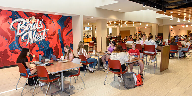 Busy lounge with people seated at tables, a mural saying 'The Bird's Nest' on the wall.